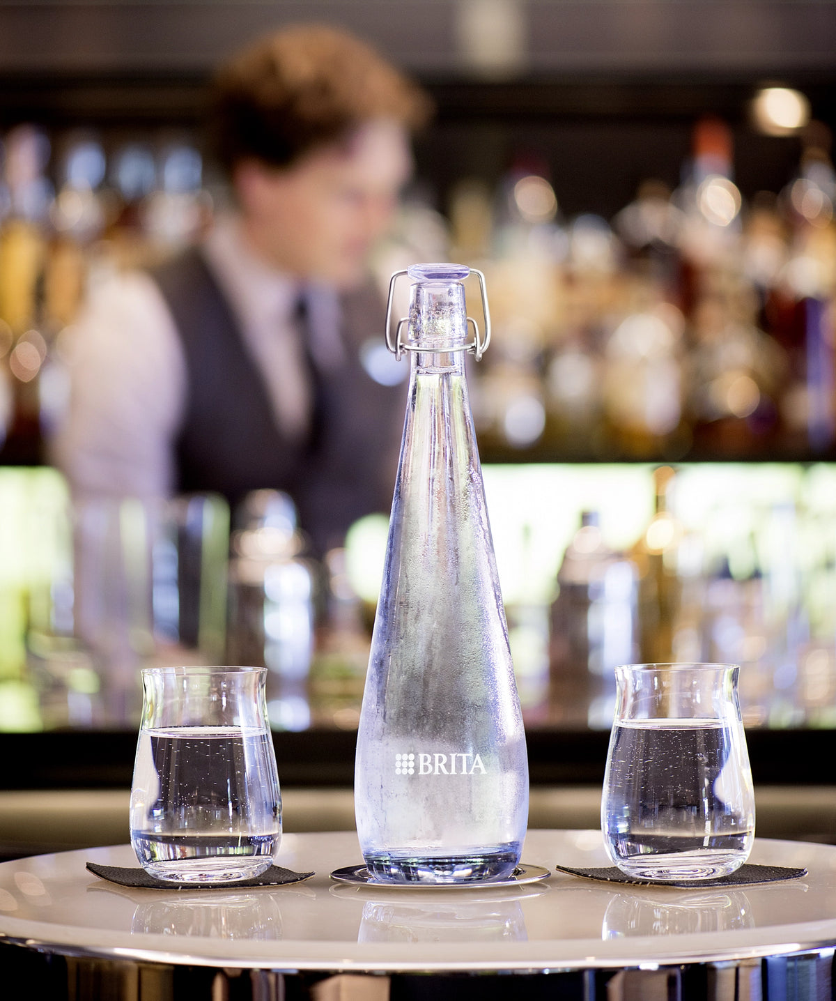 BRITA Swing bottle with two glasses of water on a bar counter with a blurred bartender in the background.