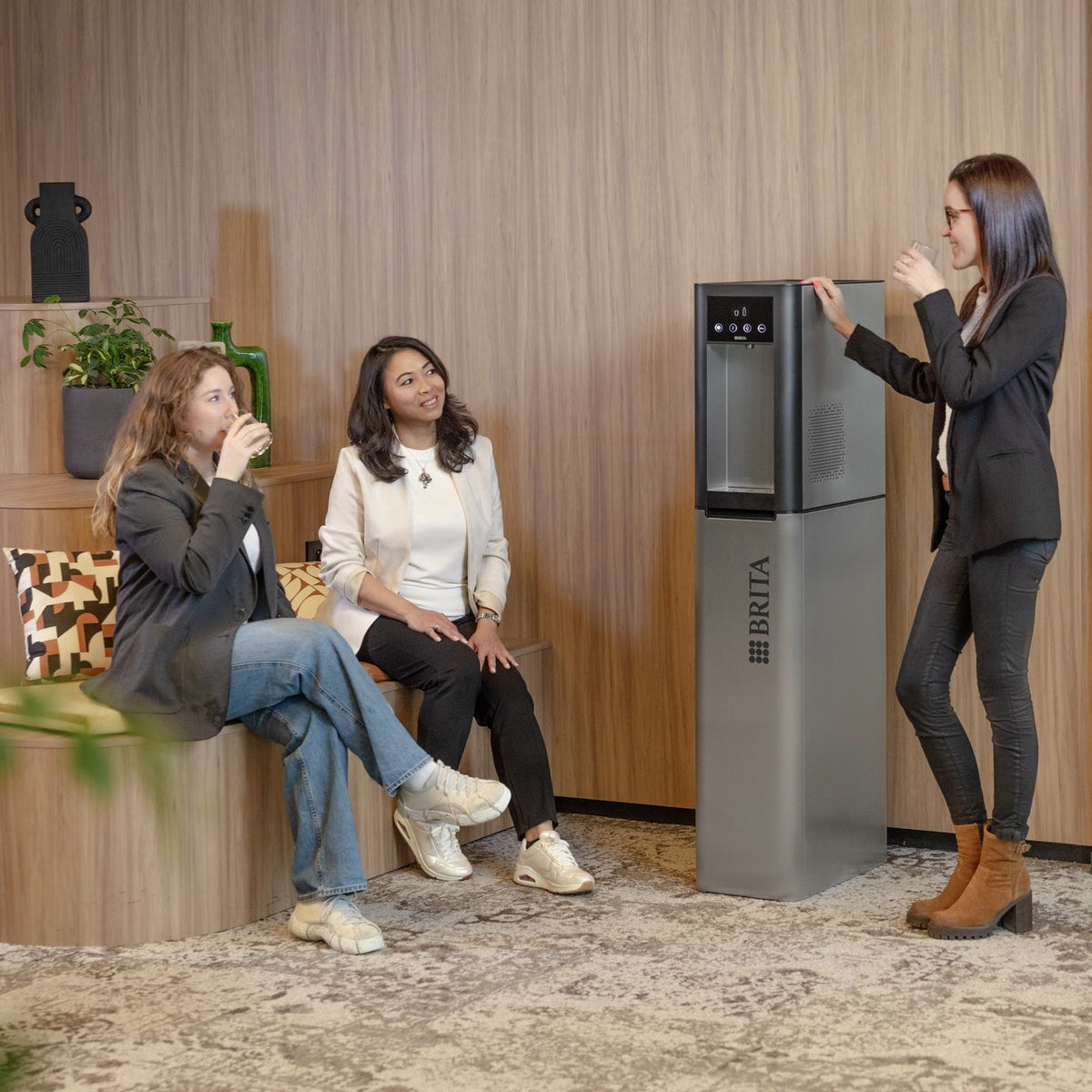 Woman standing next to a Top Pro water dispenser in a modern office setting with two other women sitting on a bench.